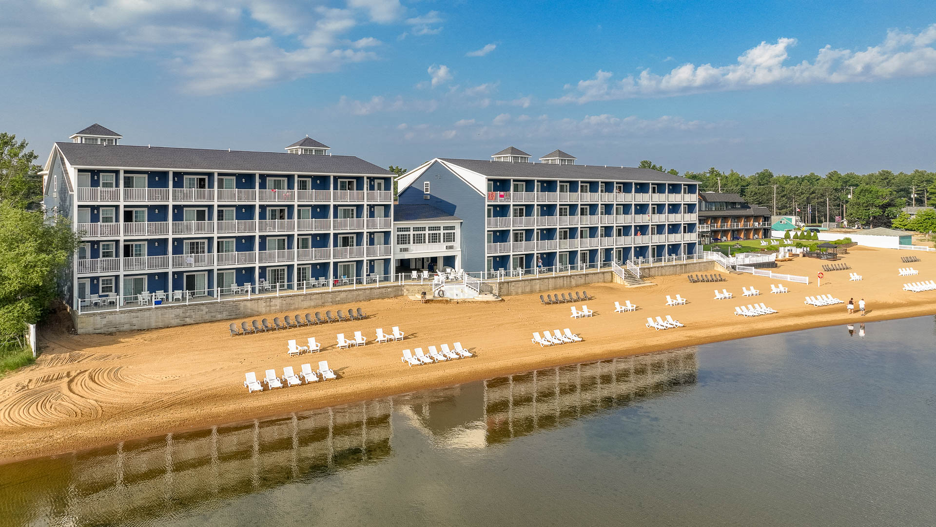 A large blue and white building with balconies stands on a beach next to calm water.