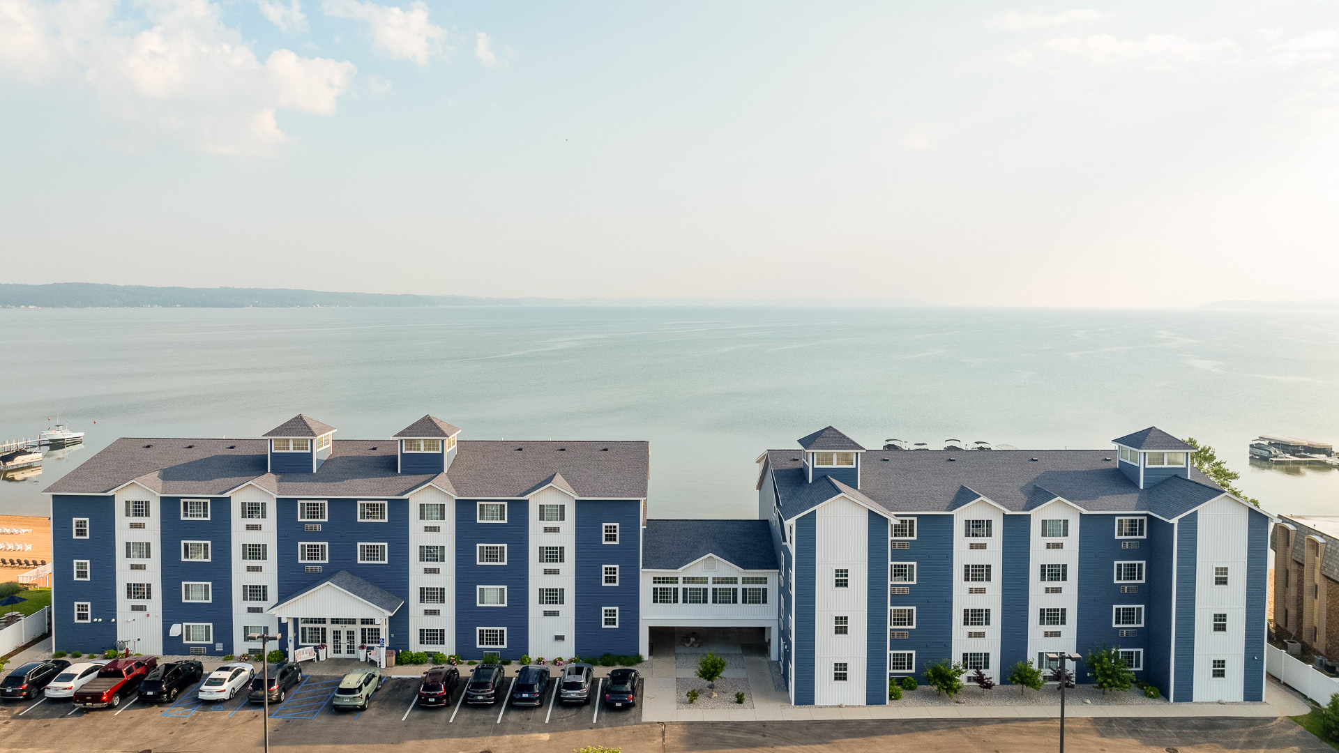 Aerial view of a blue and white multi-story building with a parking lot in front of it, overlooking a body of water.