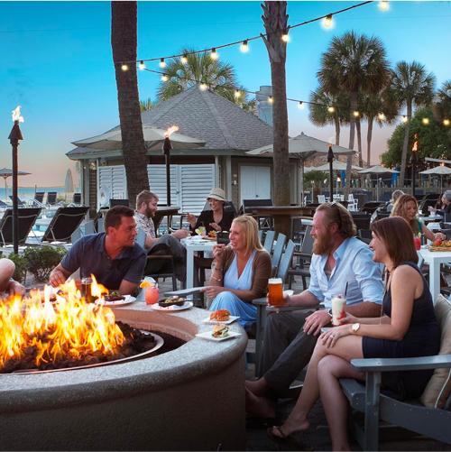 a group of friends sitting next to a fire with drinks in their hands