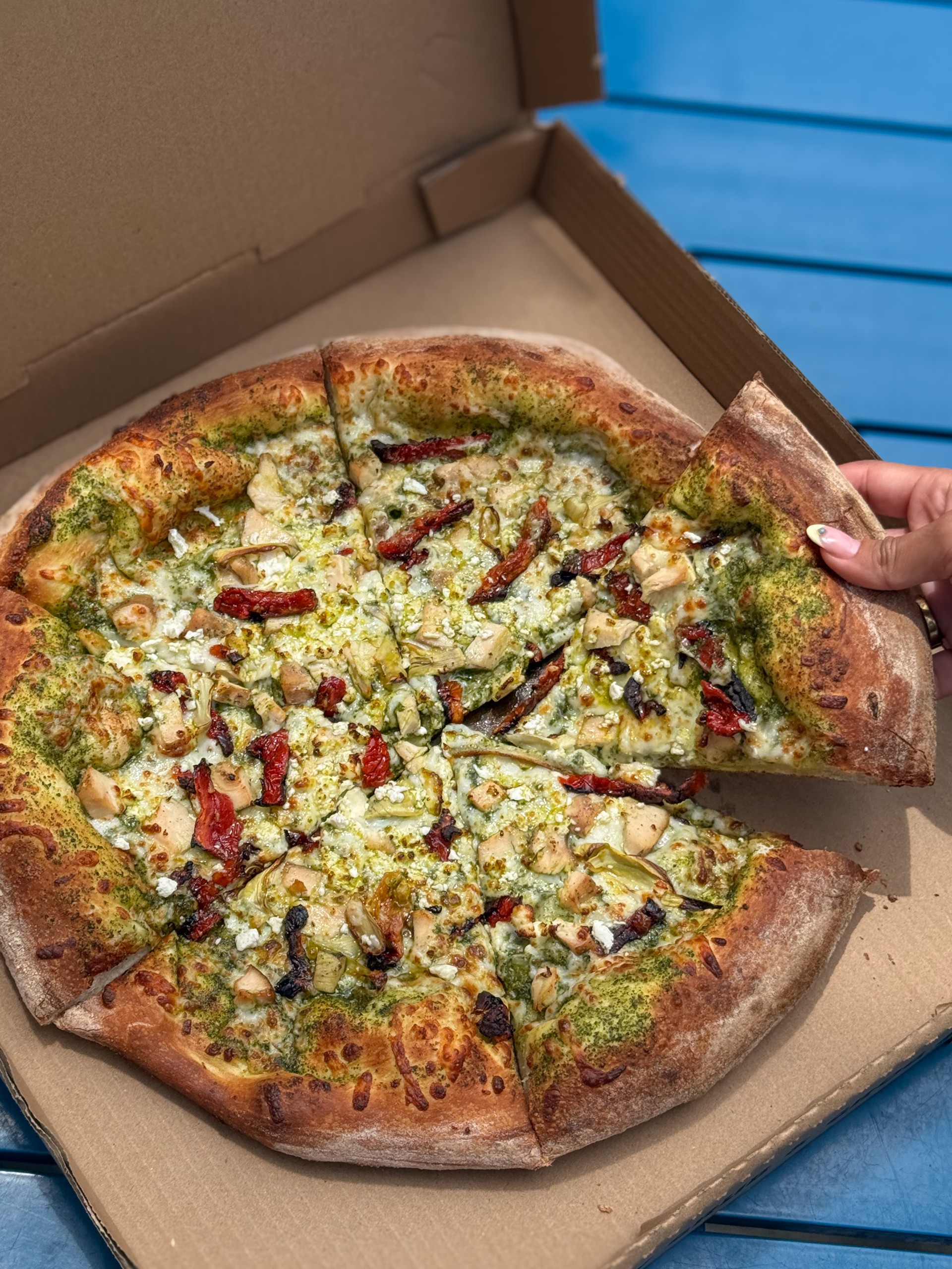 a hand holding a pesto and feta-topped pizza in a box