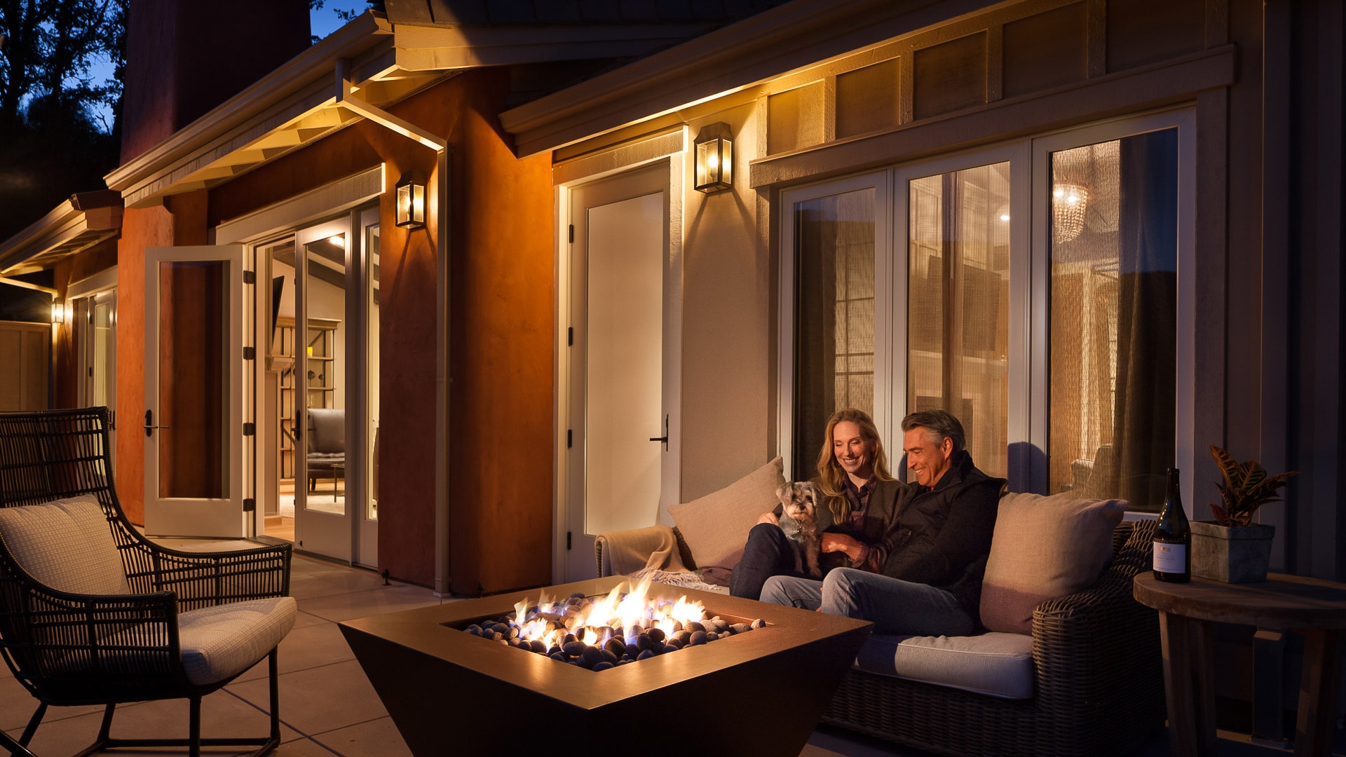 a man and woman sitting on a couch with a dog in front of a fire pit
