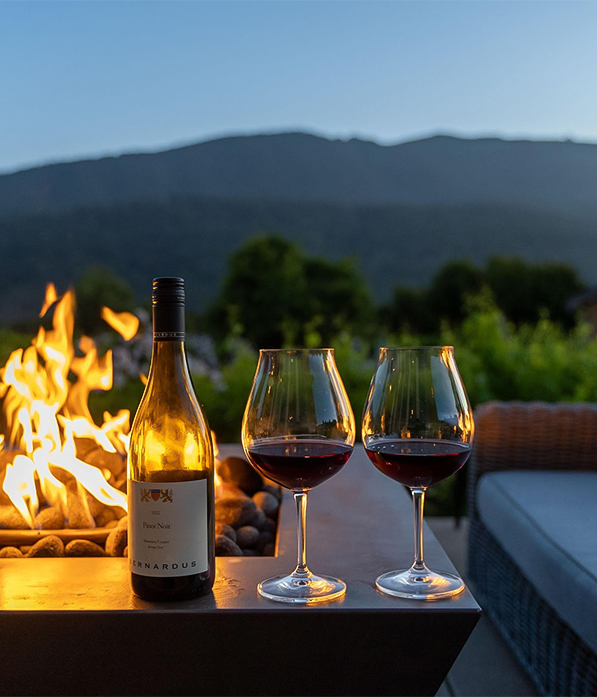 a bottle of wine and glasses on a table with a fire pit in the background