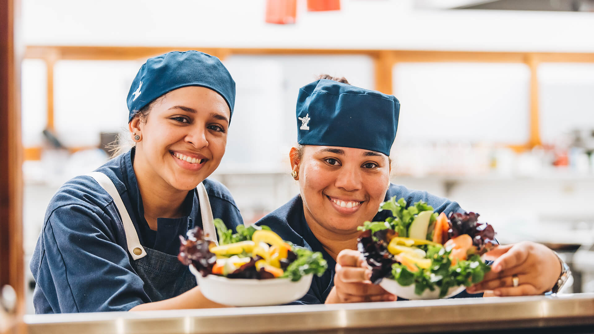 two women wearing blue hats and aprons holding a bowl of salad