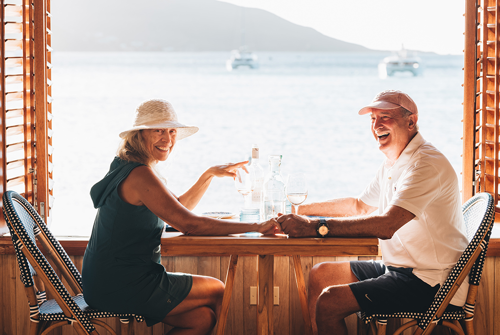 a man and woman sitting at a table with wine glasses and a body of water