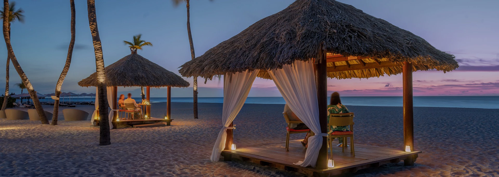 a couple of people sitting under a straw hut on a beach