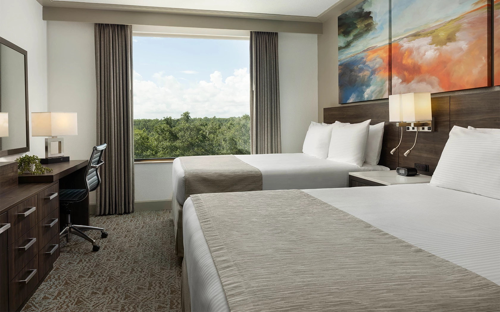A hotel room with two beds, a desk, and a window showing trees outside.