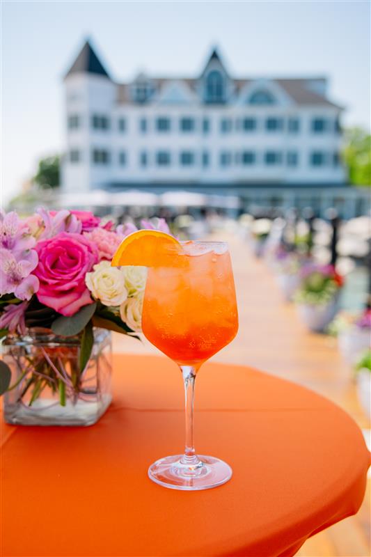 a glass of orange liquid with a slice of orange on a table with flowers in a vase