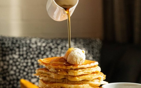Hand pouring syrup on a stack of buttermilk pancakes with a cup of coffee