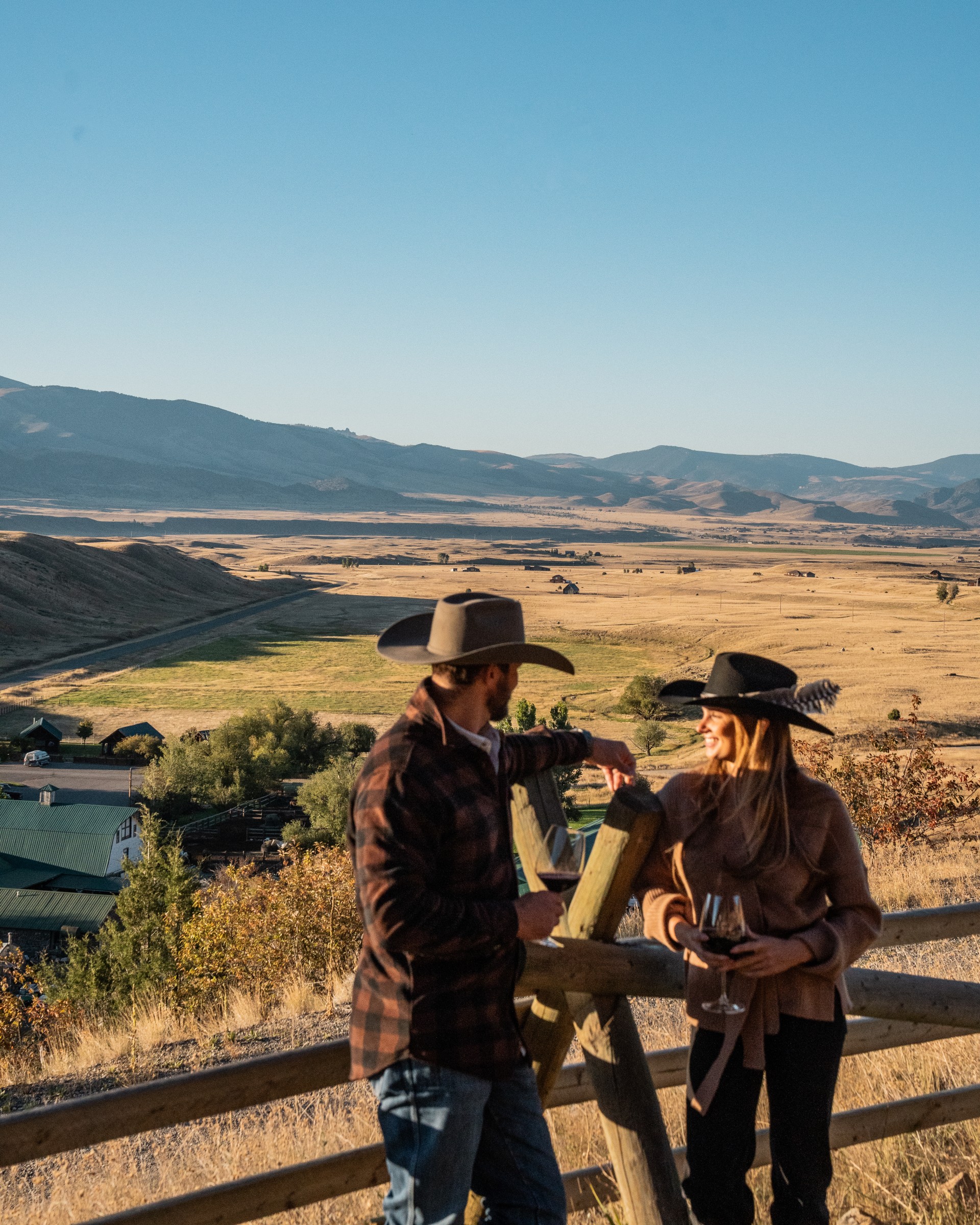 a man and woman in cowboy hats standing on a fence with a field and mountains in the background