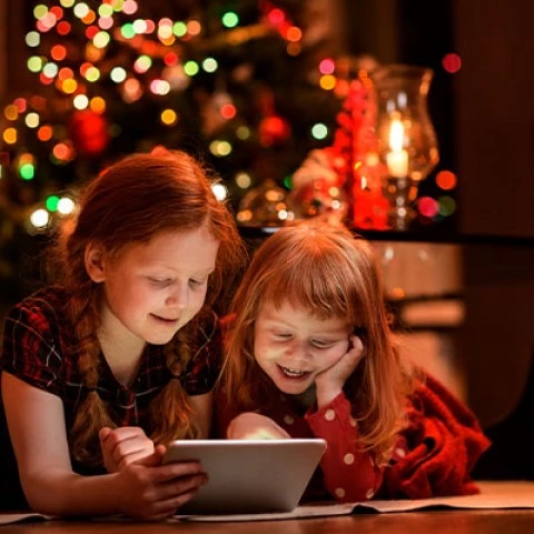 two young girls smiling and looking at a tablet in front of a christmas tree