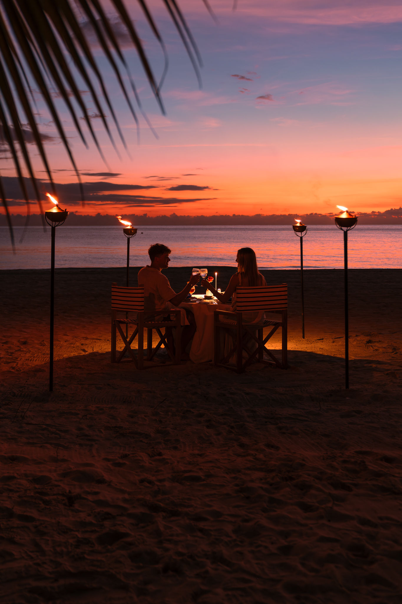 a man and woman sitting at a table on a beach with torches