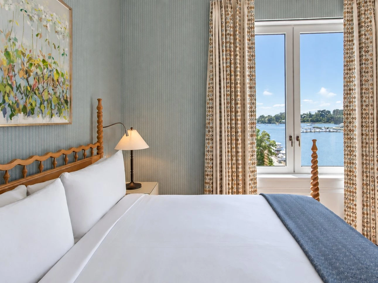 A neatly made bed with white linens and a blue throw blanket, facing a window with a view of the water.