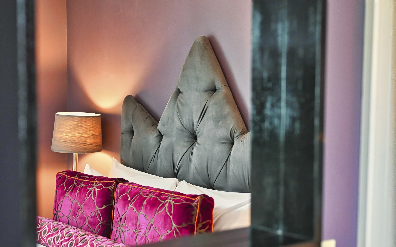 A close-up of a bed with a tufted headboard, velvet pillows, and a bedside lamp, against a purple wall.