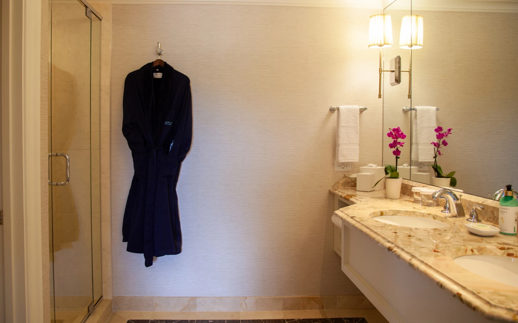 A luxurious bathroom with a marble countertop, double vanity, and a shower with a glass door. A bathrobe hangs on the wall next to the shower.