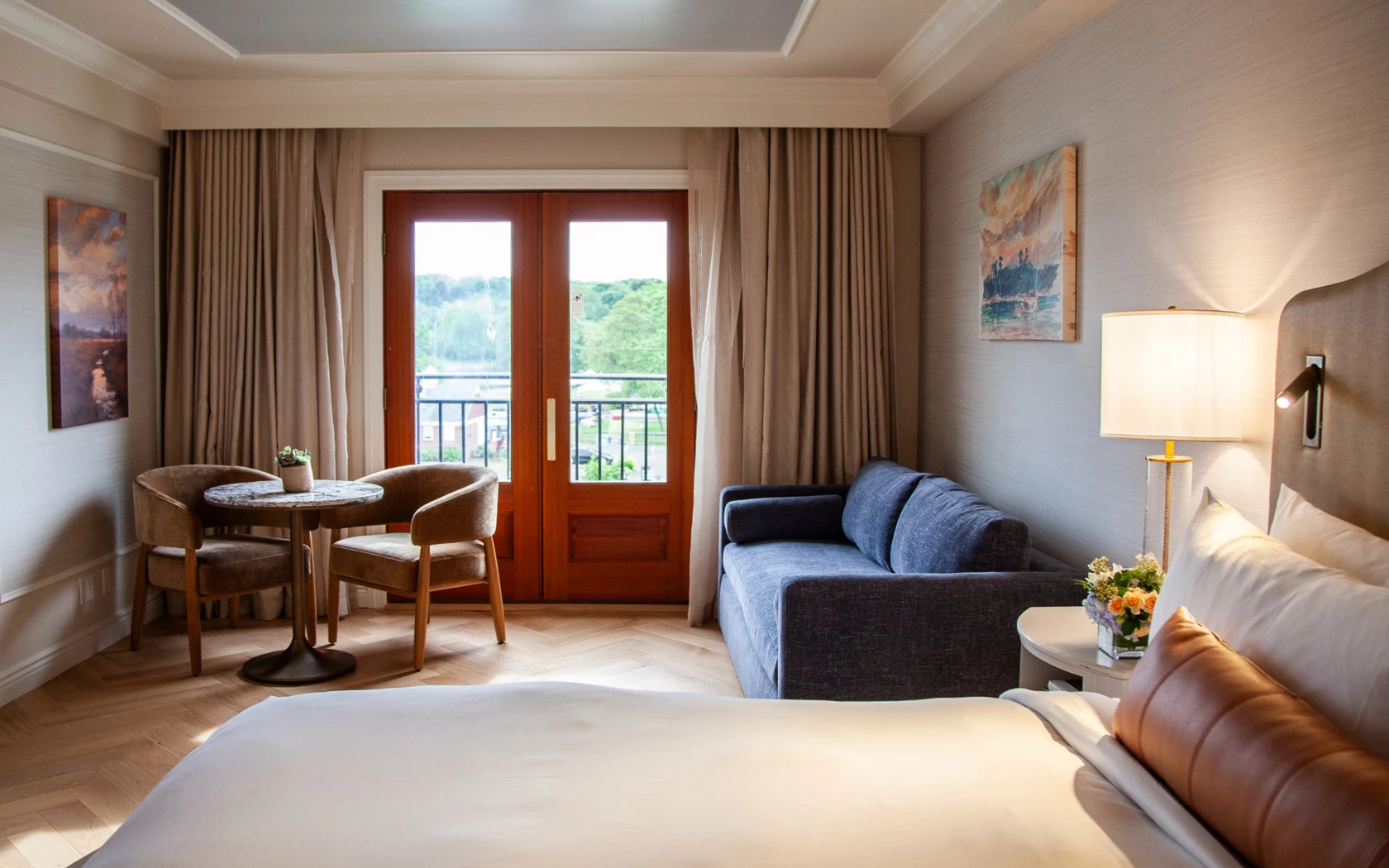 A hotel room with a king-sized bed, a sofa, a table with two chairs, and a balcony door with a view.