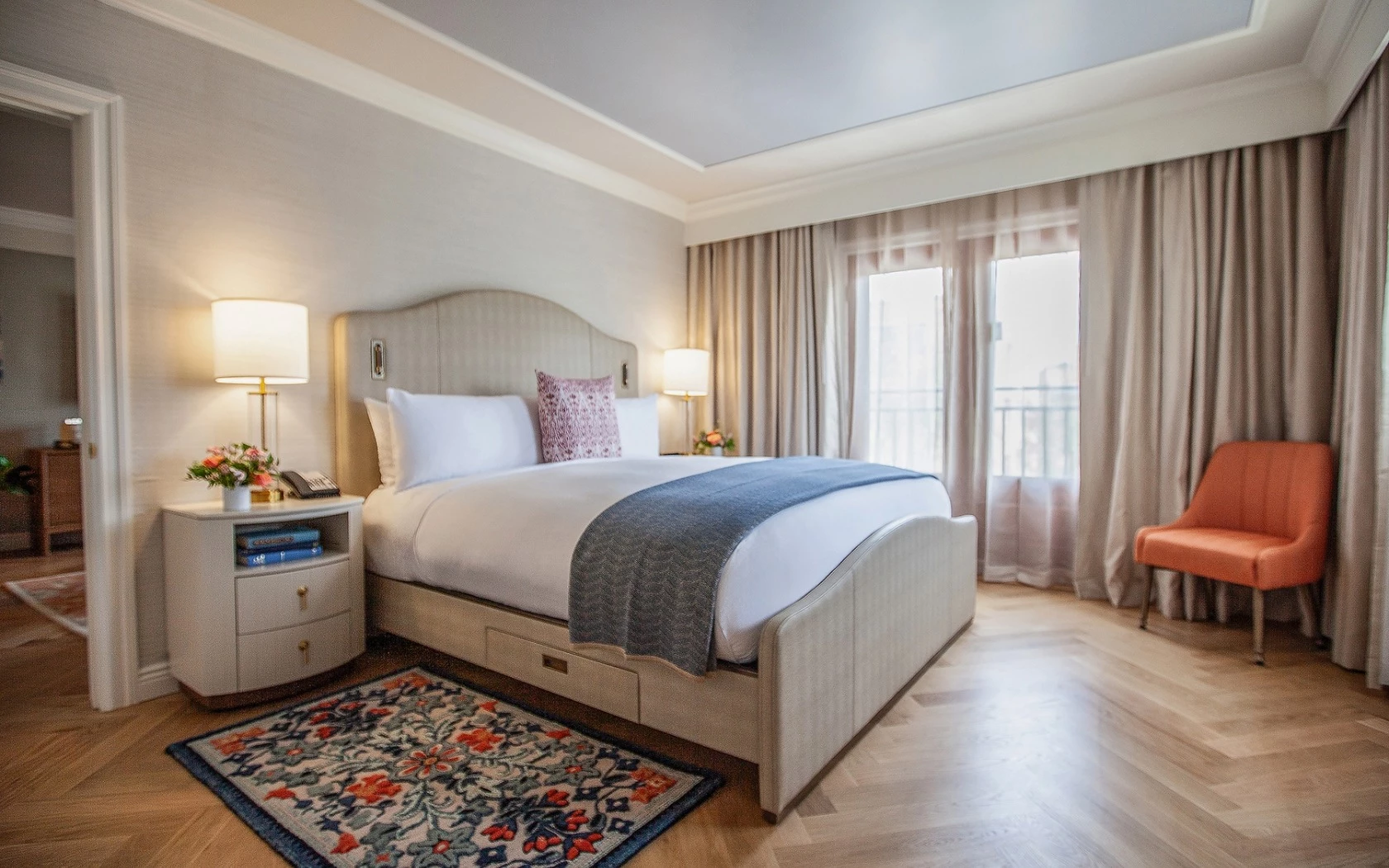 A hotel room with a bed, nightstand, lamp, curtain, chair, and rug.