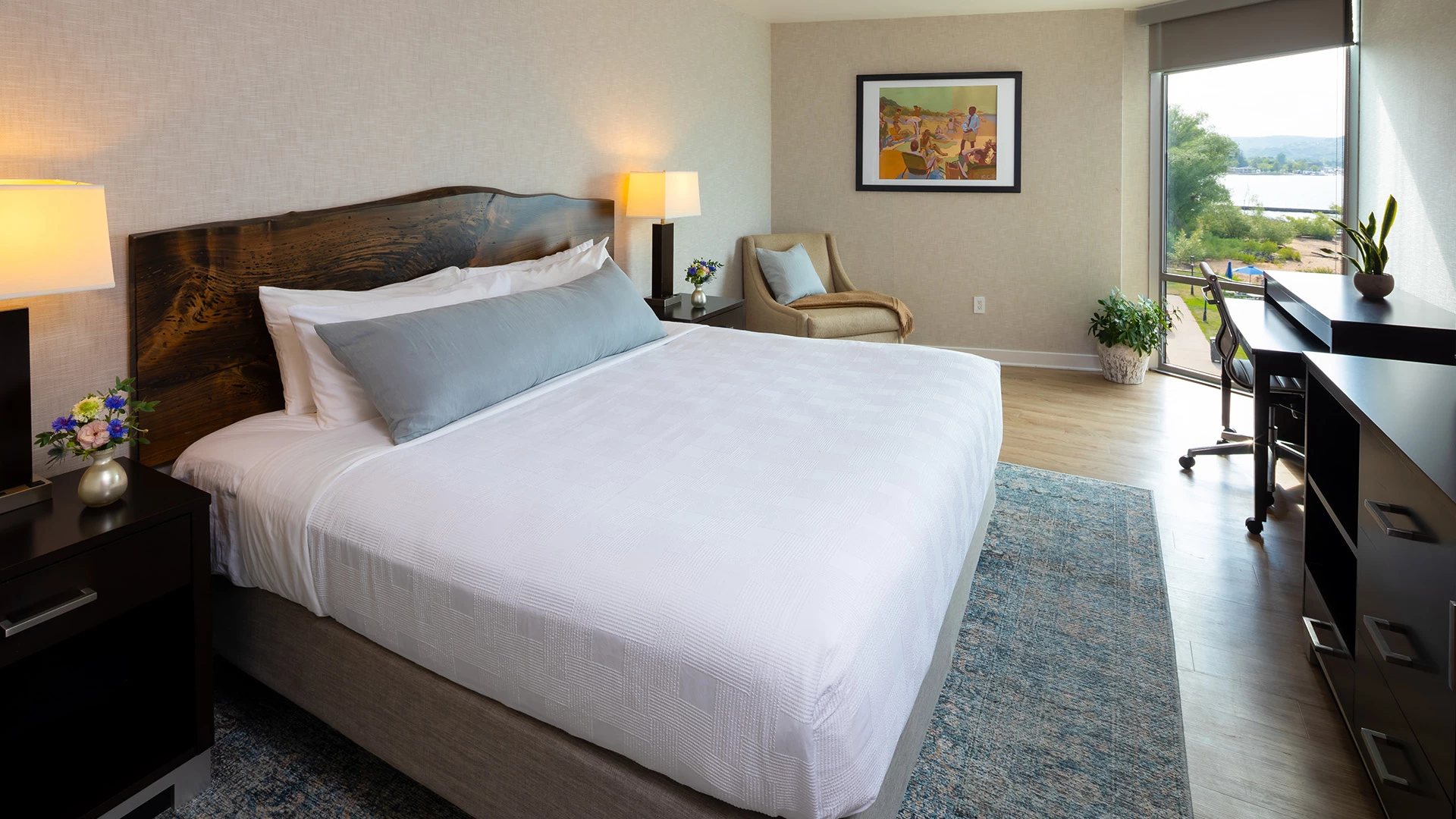 A wide view of a hotel room with a large bed, nightstands, a chair, and a desk in front of a window with a view.