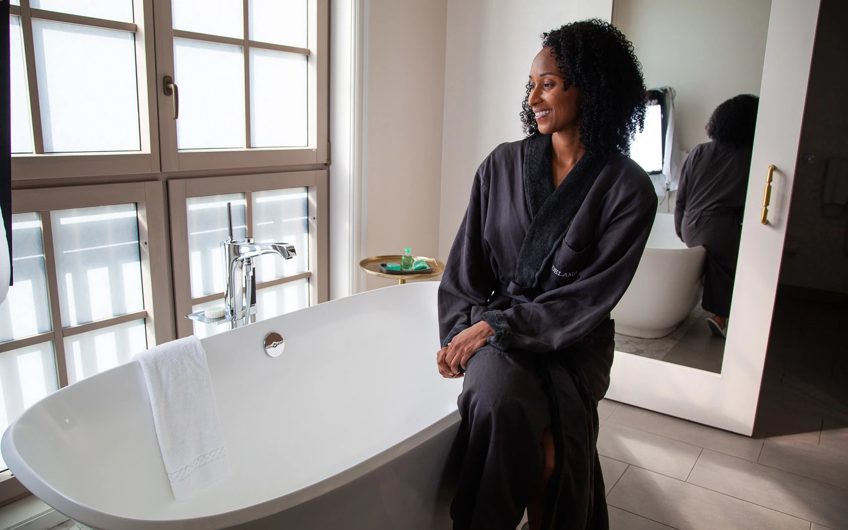 A woman in a black robe sitting on a freestanding tub in front of a large window with a tray and mirror nearby.
