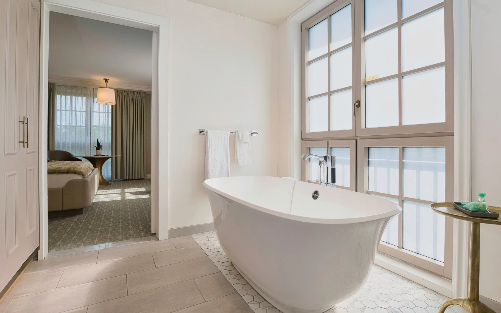 A large white soaking tub sits in front of a window in a modern bathroom with a tiled floor and towels hanging on the wall.