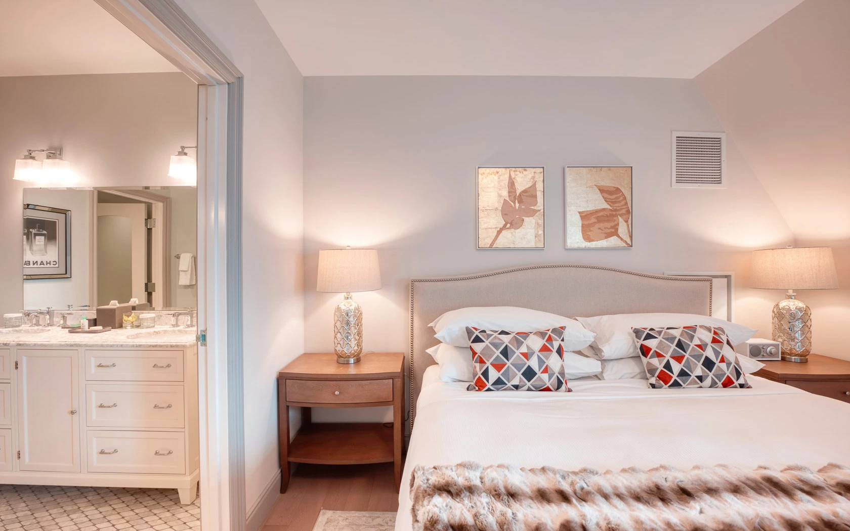 A neatly made bed with pillows and a fur throw in a bedroom with a bathroom in the background.