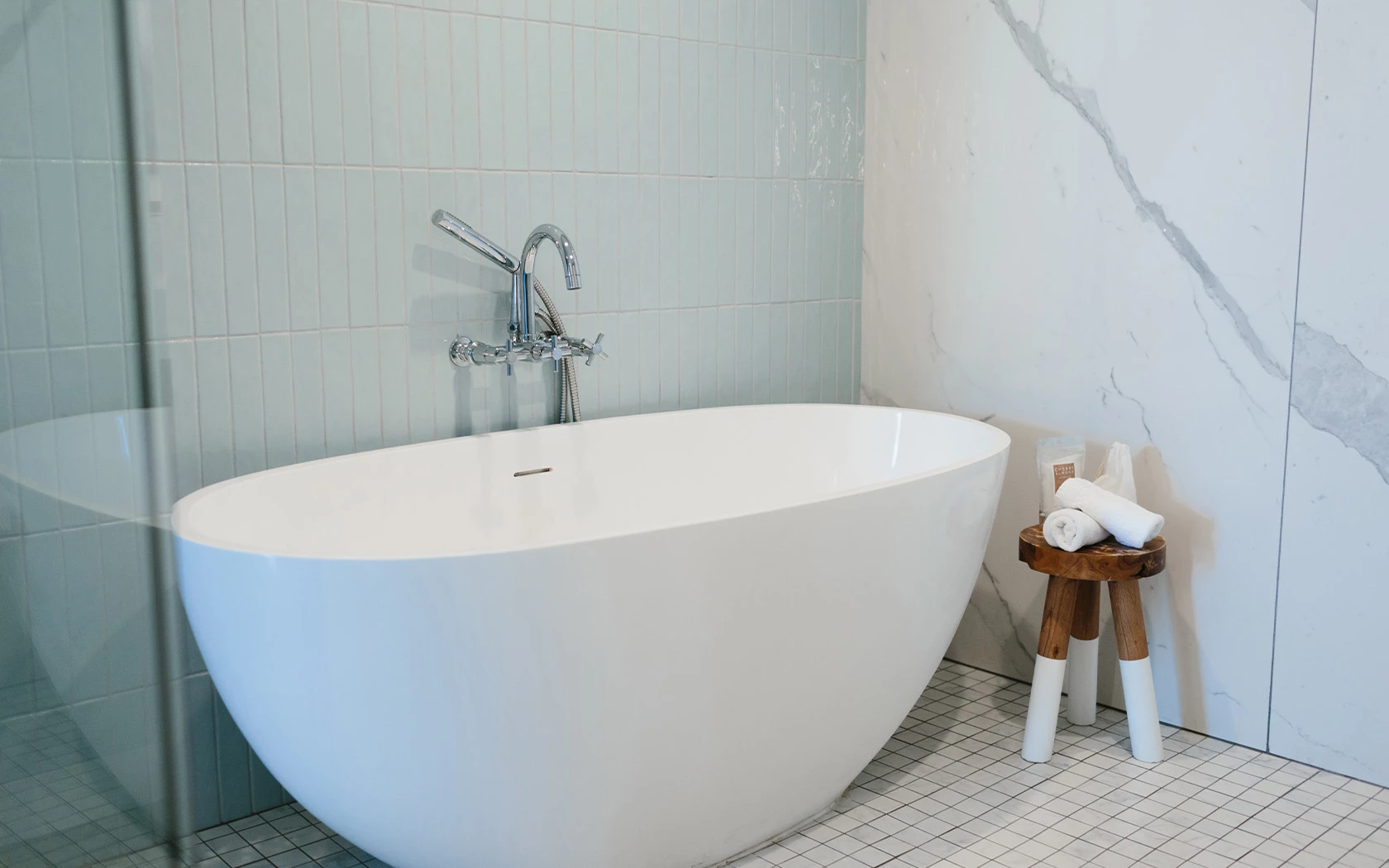 A white bathtub with a silver faucet, tiled walls, and a tiled floor, with a small wooden stool holding two rolled towels.
