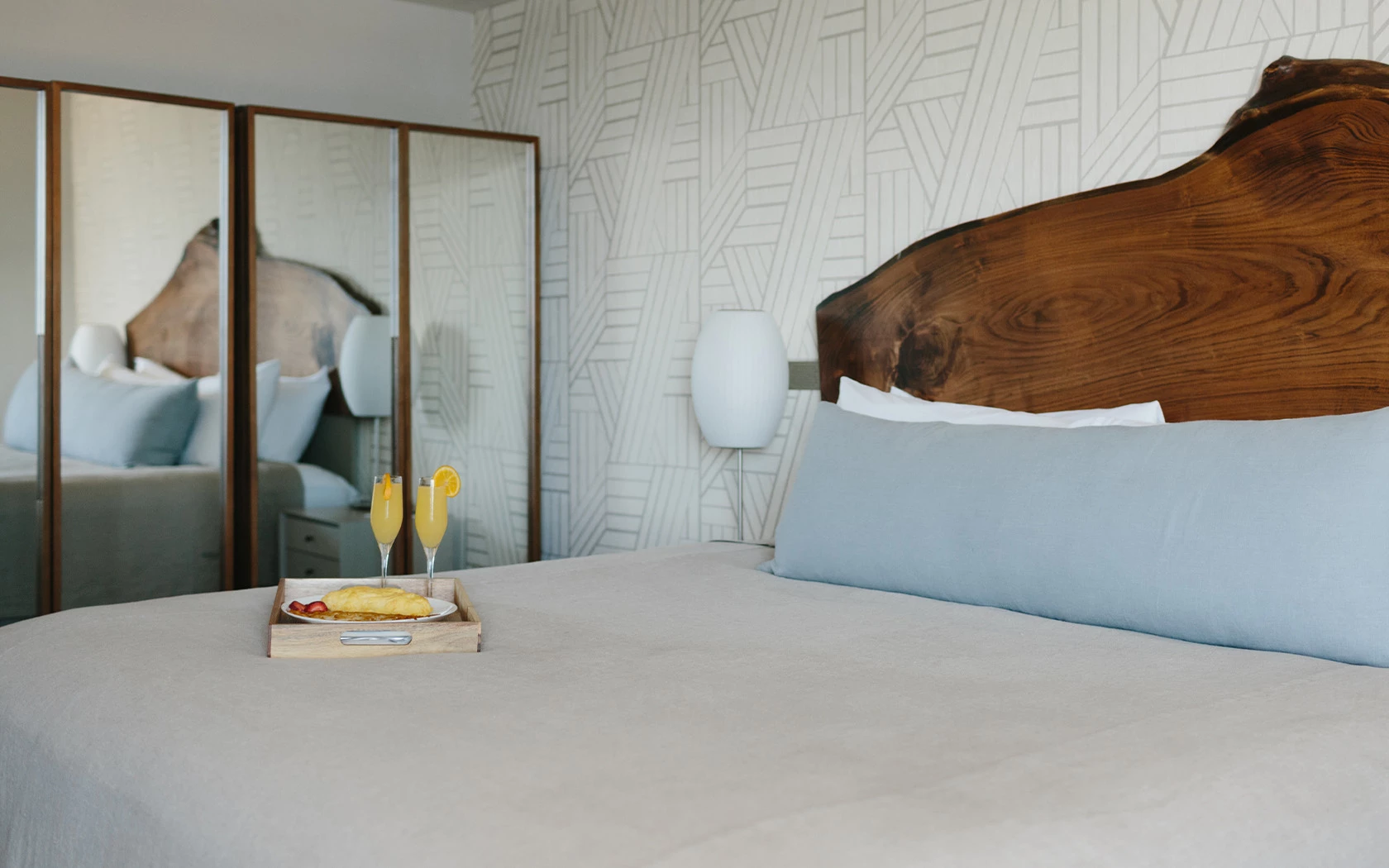 A close-up of a bed with a wooden headboard, light blue pillows, and a white comforter, with a tray of food and drinks on it.