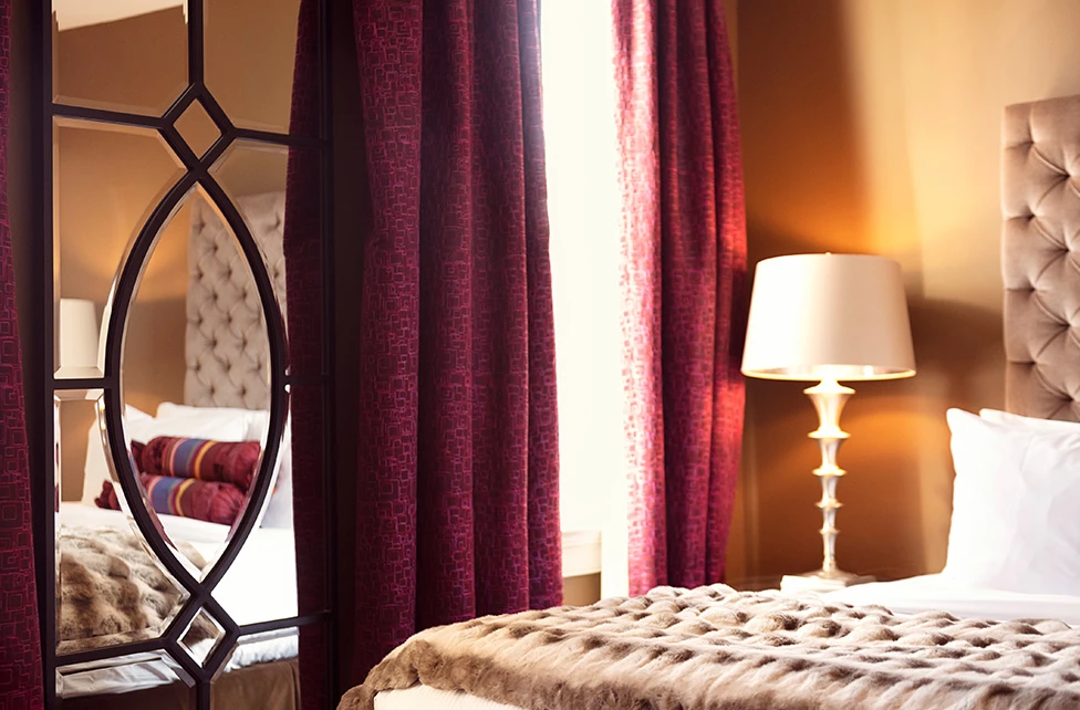 A close-up view of a bed with a white comforter and pillows, accompanied by a lamp and a mirror in a room with red curtains.