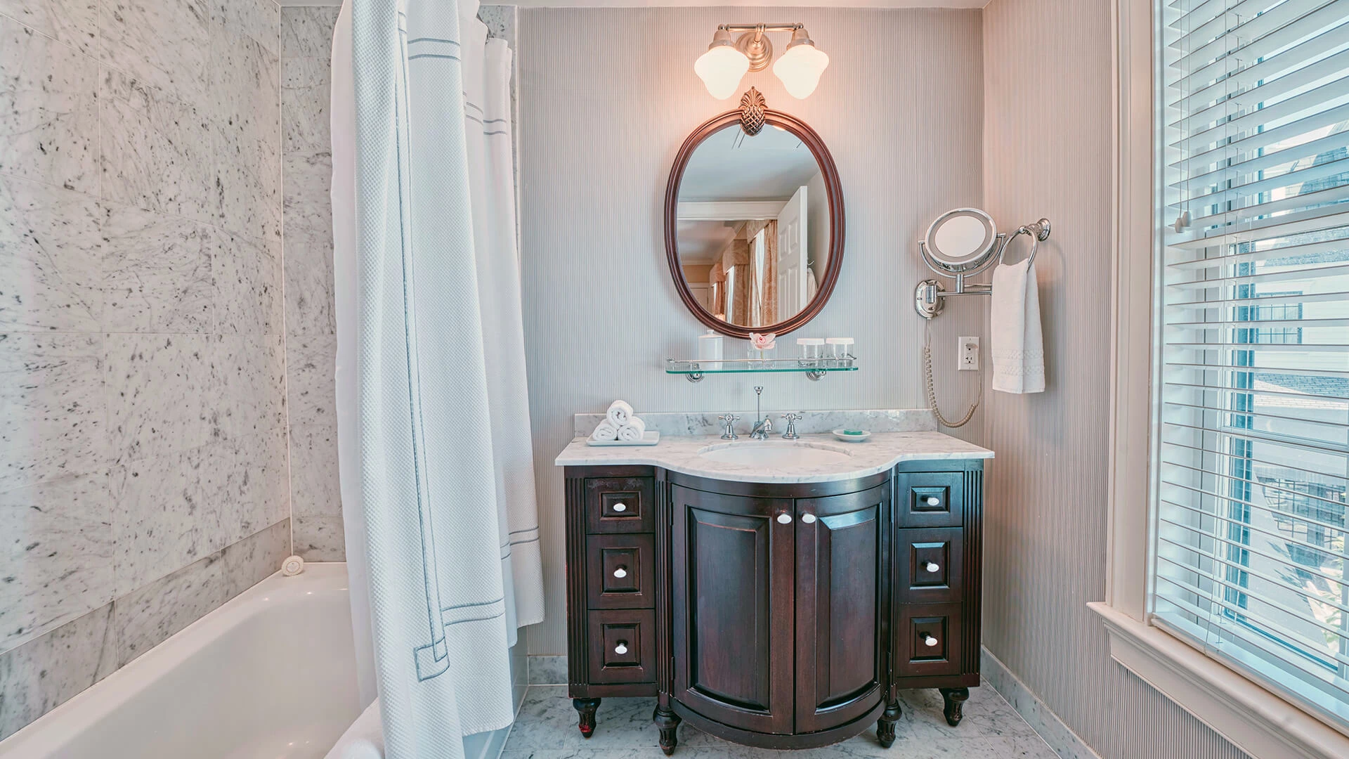 A bathroom with a marble vanity, bathtub, and window with blinds.