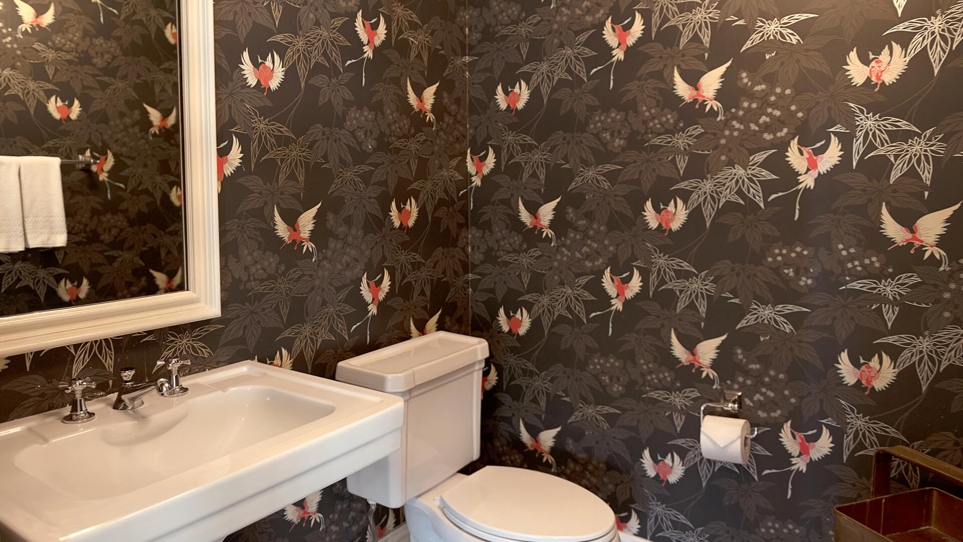 A bathroom with a toilet, sink, and mirror in front of a wall with dark floral wallpaper.