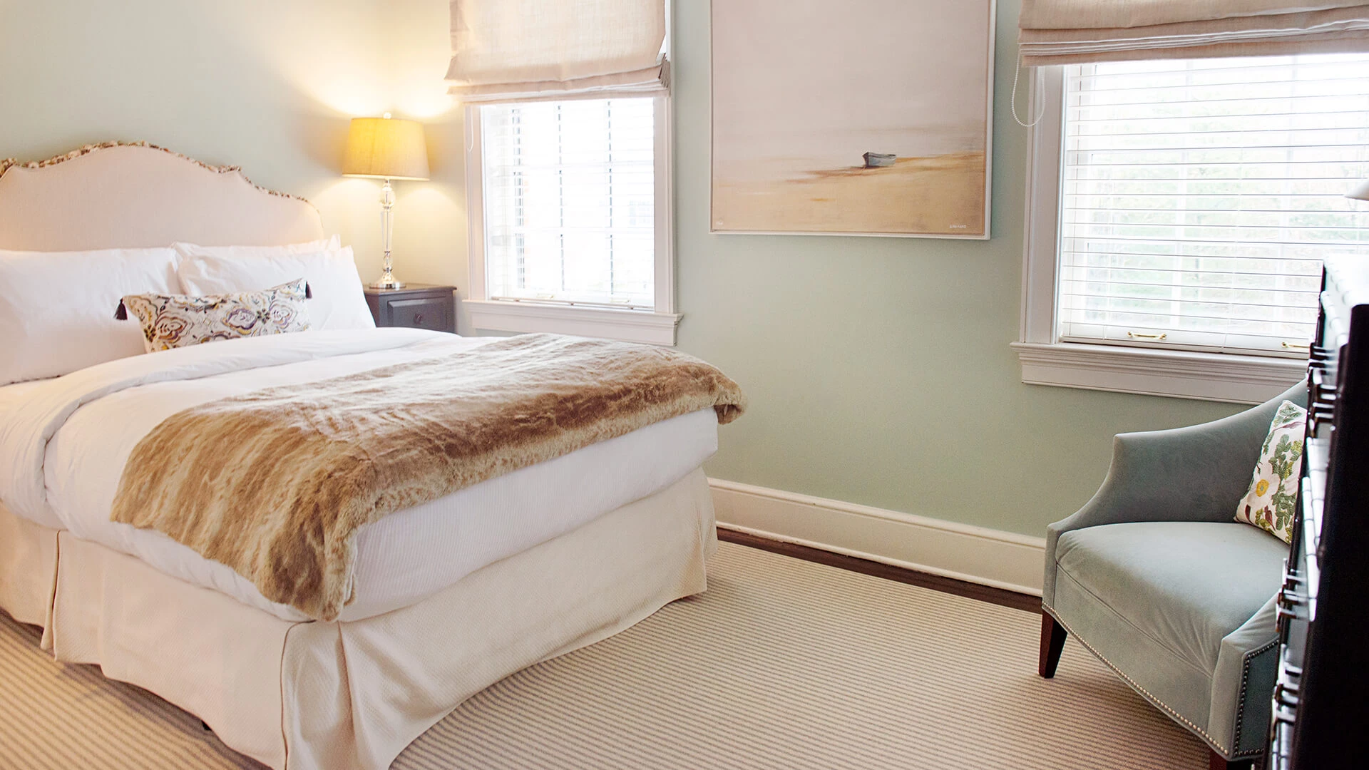 A neatly made bed with white linens and a fur throw, a chair, and two windows in a light green room.