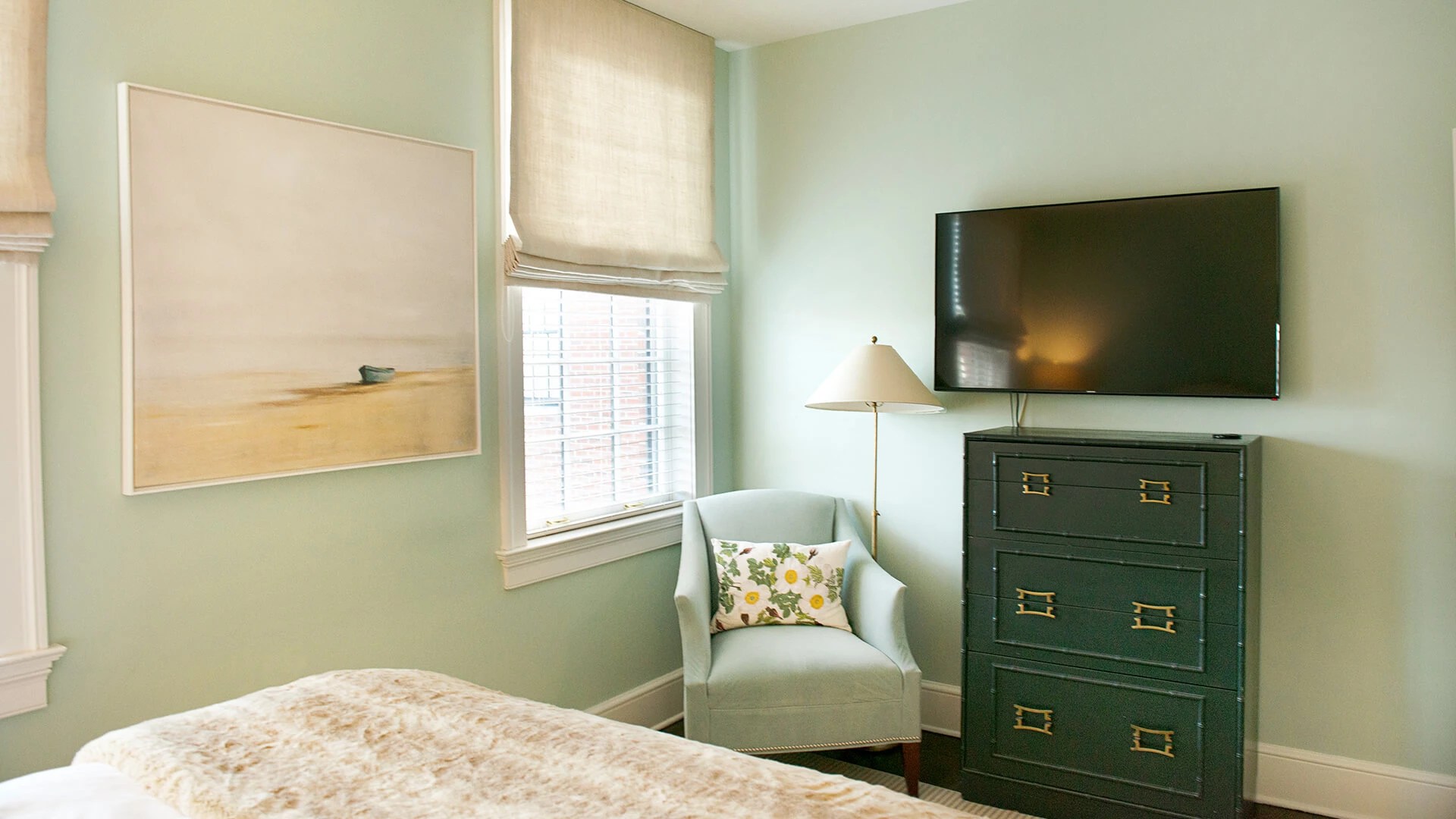 A serene bedroom with light green walls, a large window, and a comfortable bed with a floral chair and dresser.