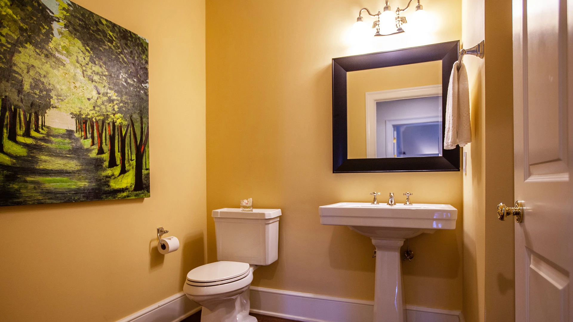 A bathroom with a toilet, sink, mirror, and painting on the wall.