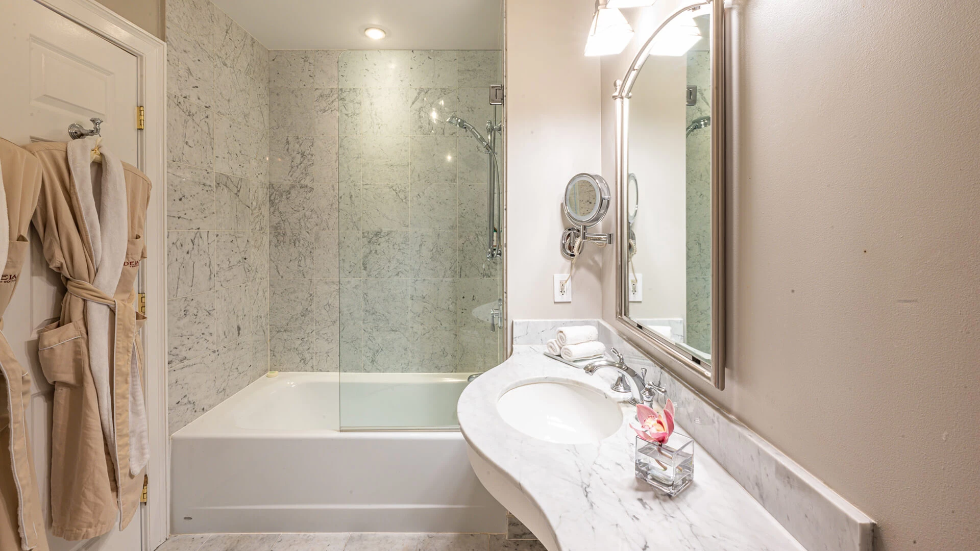 A bathroom with a marble shower and bathtub, a sink with a mirror, and robes and towels hanging on the door.