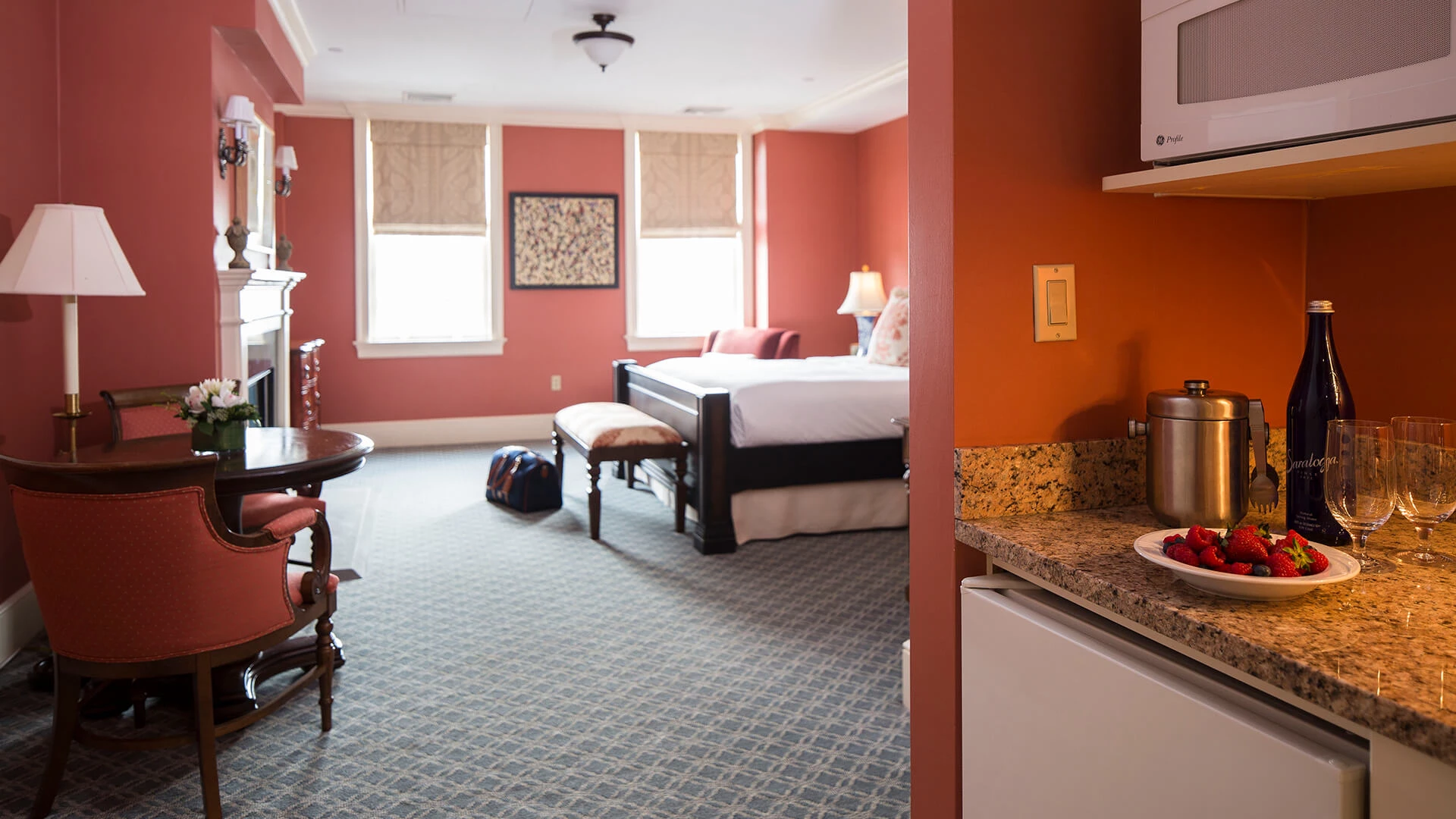 A wide view of a hotel room with a king-sized bed, a dining table, a microwave, and a plate of fruit and wine glasses on the counter.