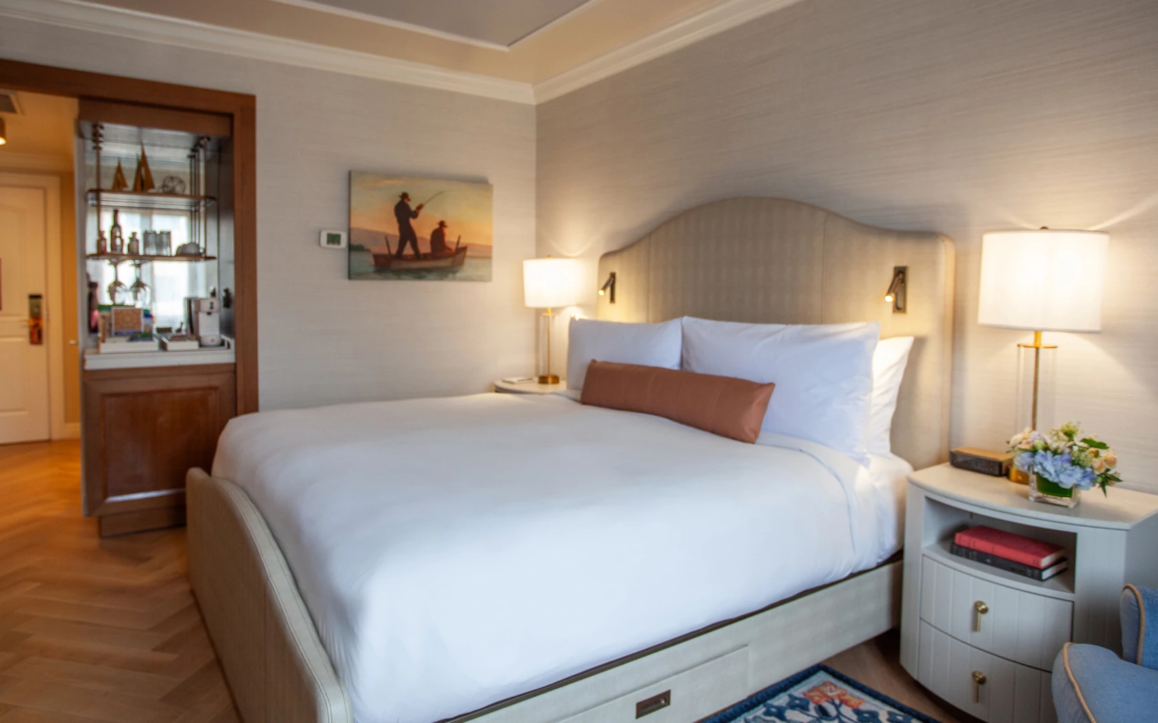 A hotel room with a large bed, two nightstands with lamps, a painting on the wall, and a bookshelf in the corner.