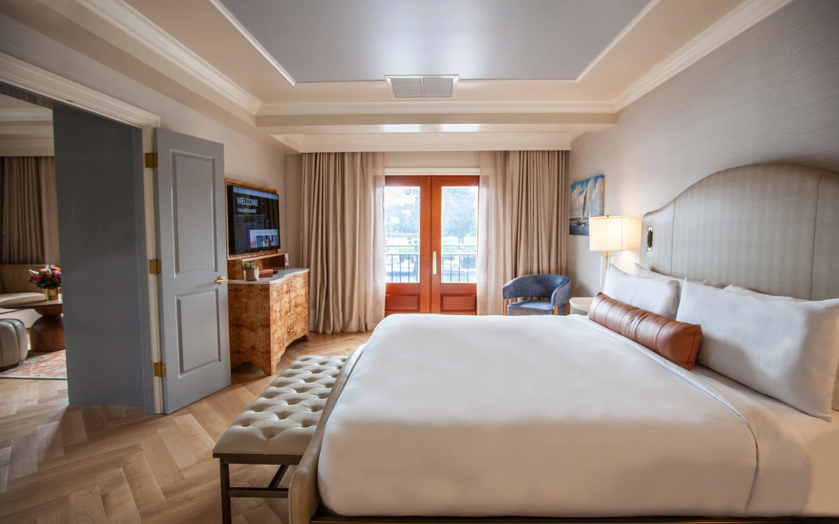 A large hotel room with a king-sized bed, wooden dresser, flat screen TV, tufted bench, and lamp.
