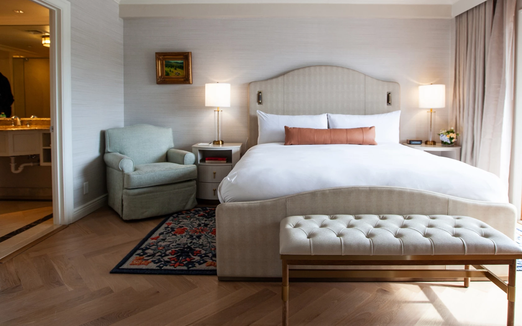 A neatly made bed with white linens and a rust-colored pillow in a room with a sitting area.