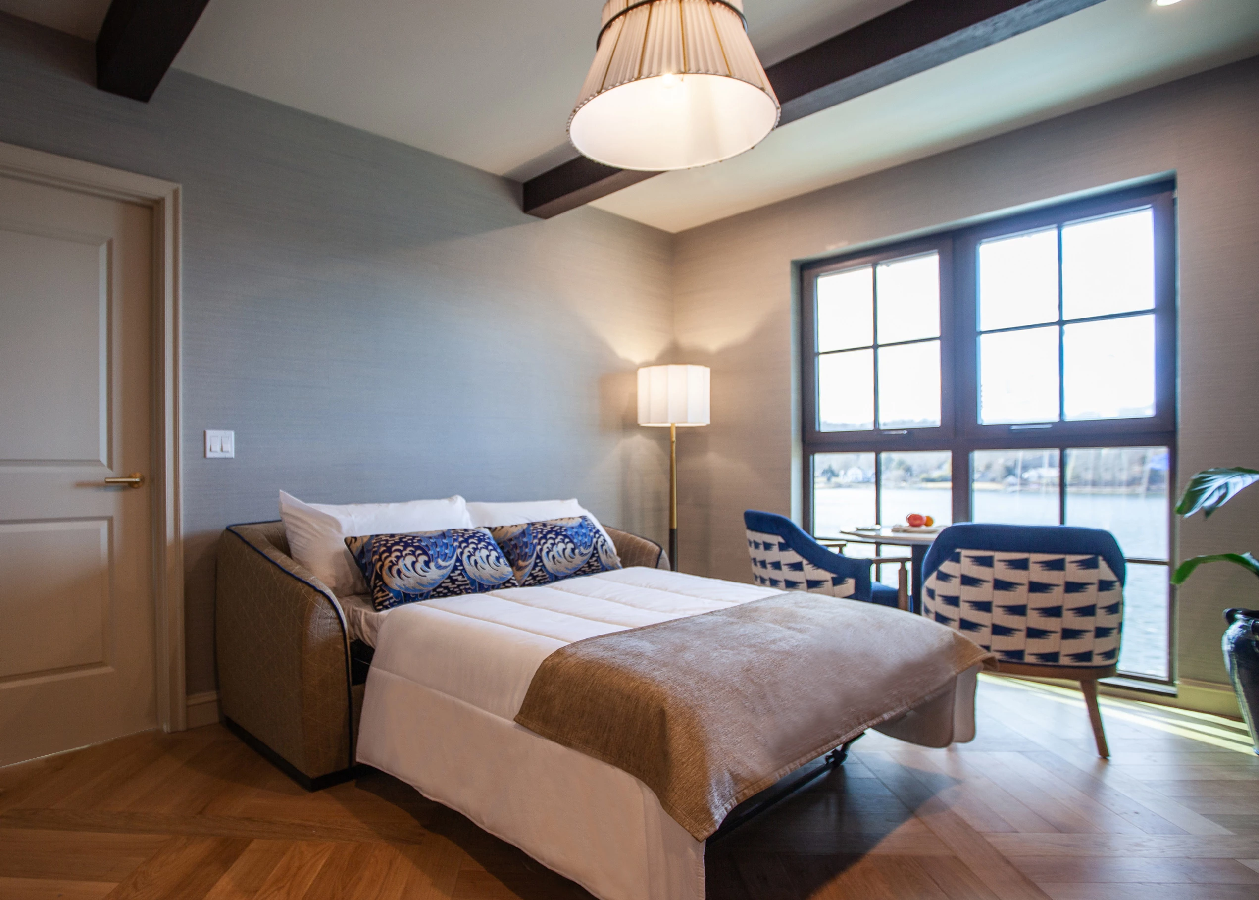A hotel room with a bed, two chairs, a lamp, and a window with a view.