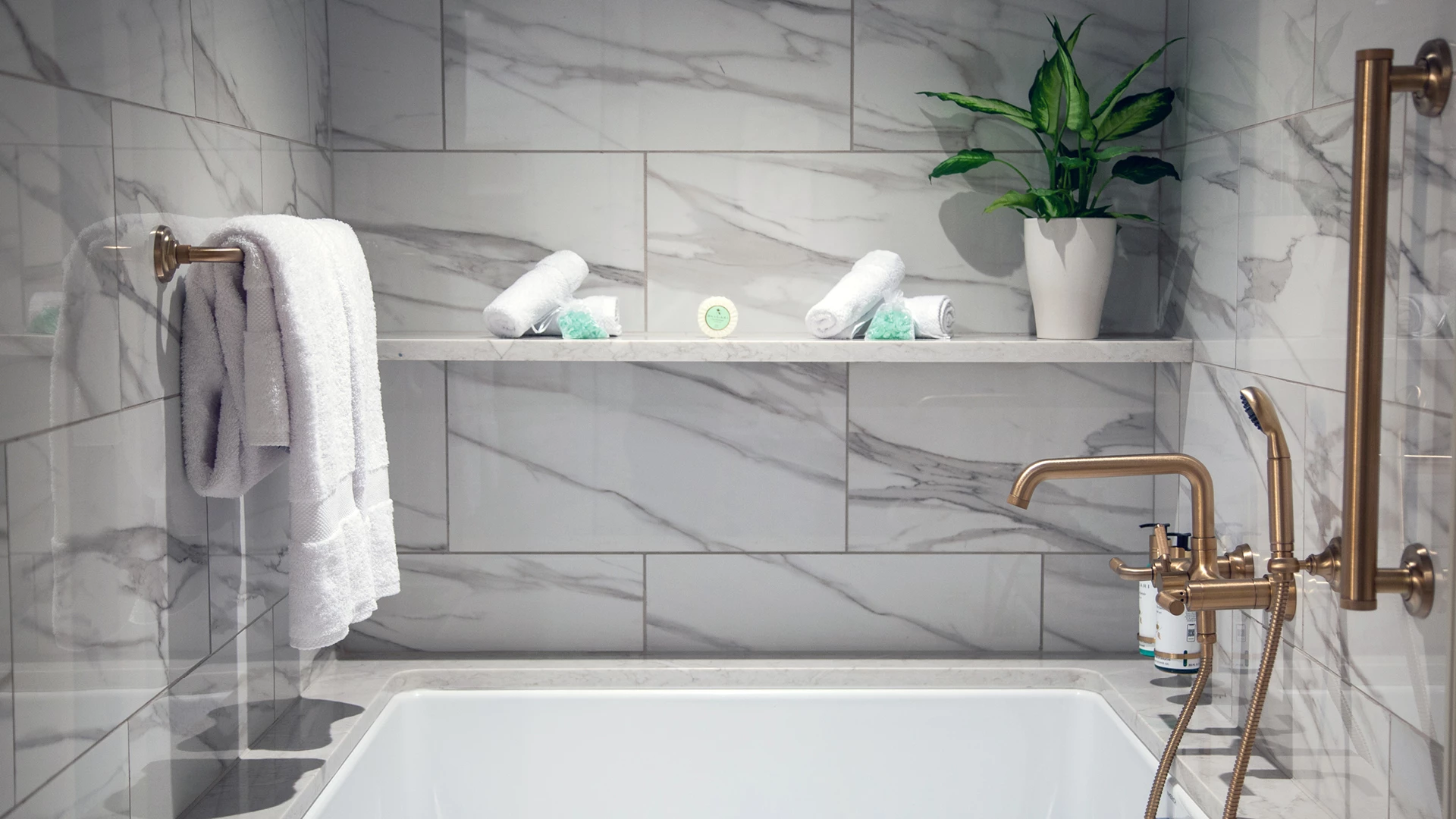 A bathroom with a white bathtub, gold faucet, and marble tile walls and floor, with towels and a plant on a shelf.