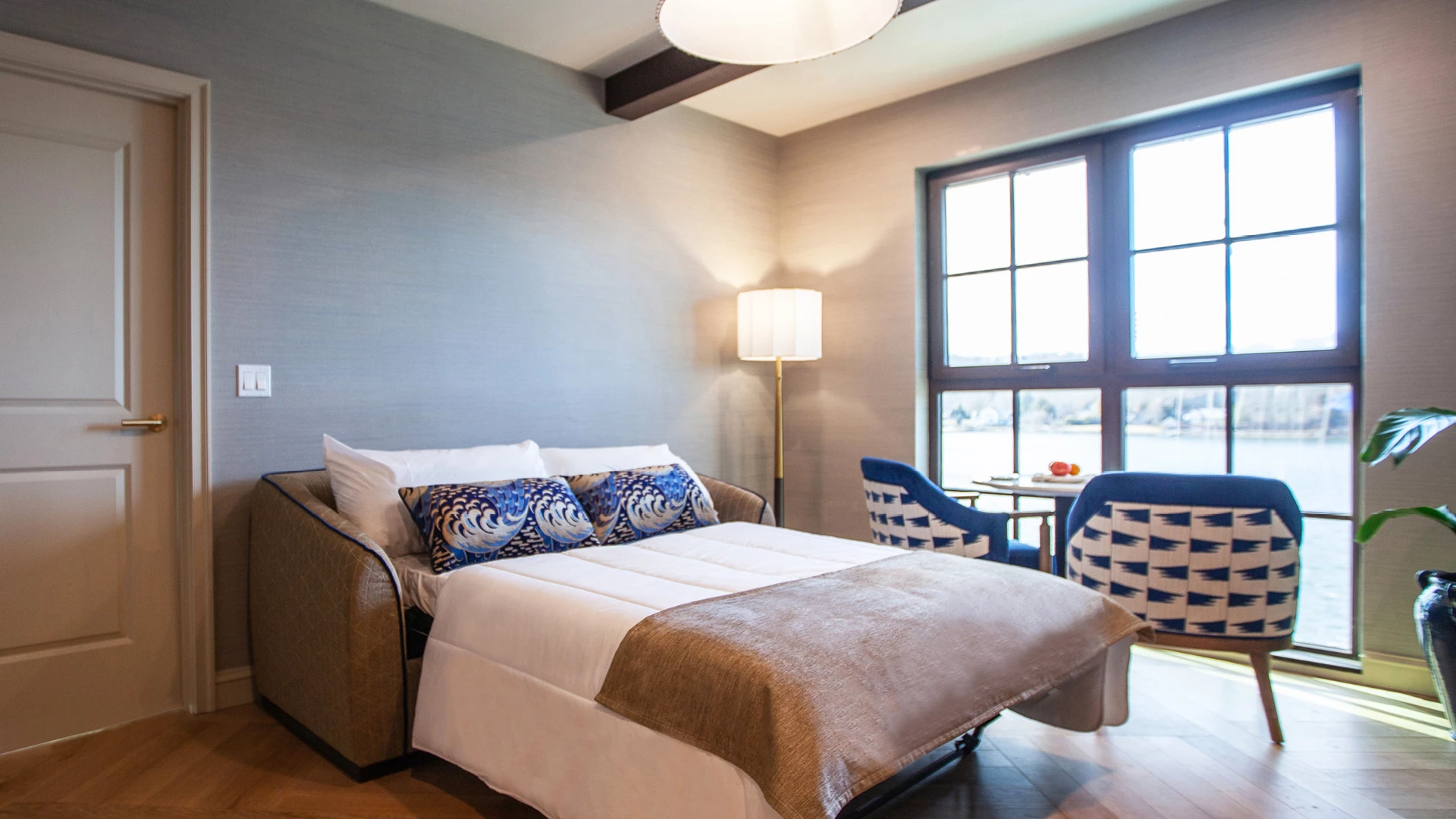 A hotel room with a bed, two chairs, a lamp, and a window.