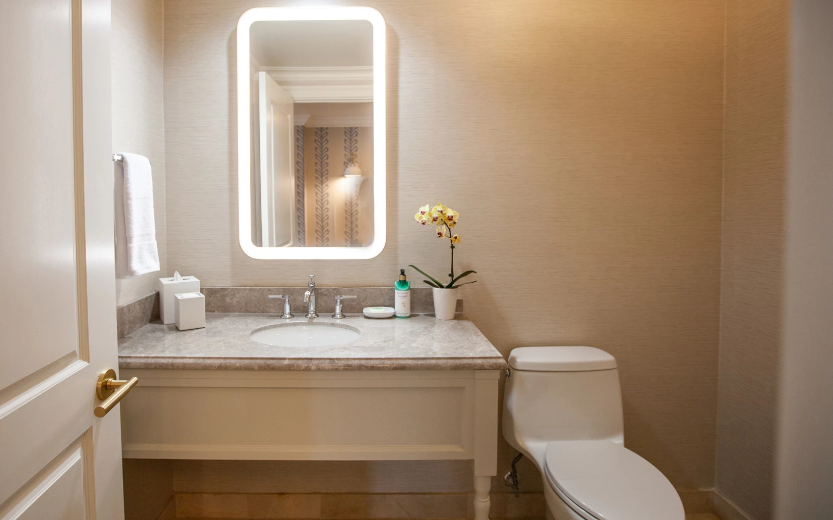 A bathroom with a marble countertop, lit mirror, toilet, sink, and towel.