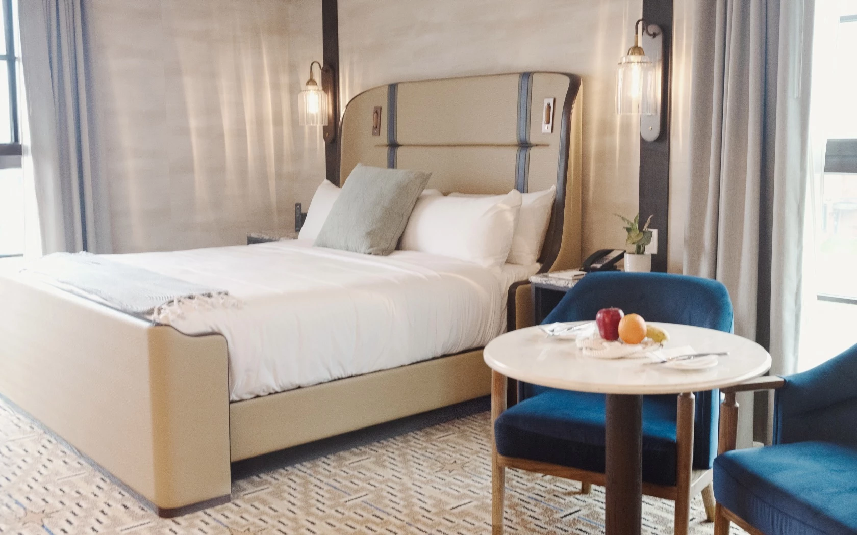 A hotel room with a bed, two blue chairs, and a small table with fruit on it.