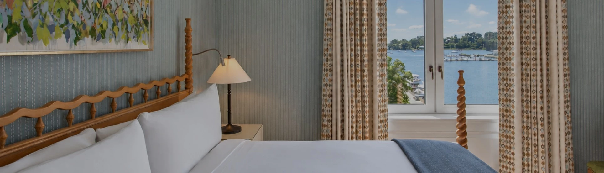A hotel room with a bed, lamp, painting, and window with a view of the water.