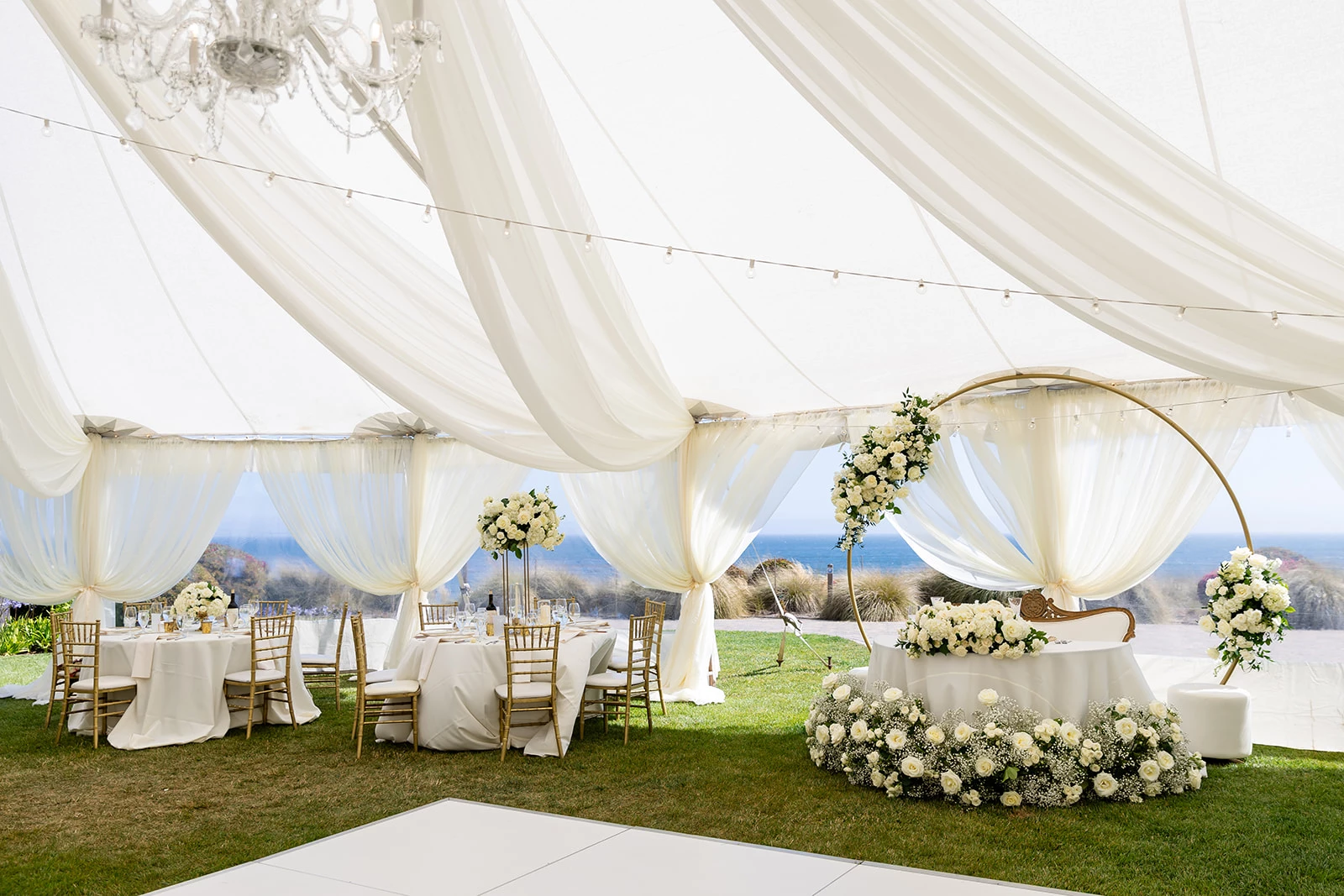 Elegant wedding reception inside the Pacific View Tent at Dolphin Bay Resort & Spa in Pismo Beach — white floral décor, chandeliers, and gold accents with ocean views creating a refined coastal ambiance.