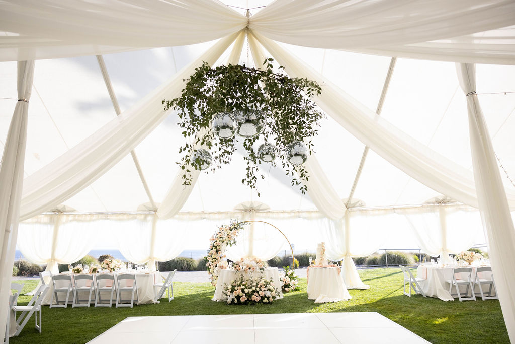 Wedding reception setup inside the Pacific View Tent at Dolphin Bay Resort & Spa in Pismo Beach — featuring white dance floor, sweetheart table, disco ball, floral chandelier, and ocean backdrop for an elegant coastal celebration.