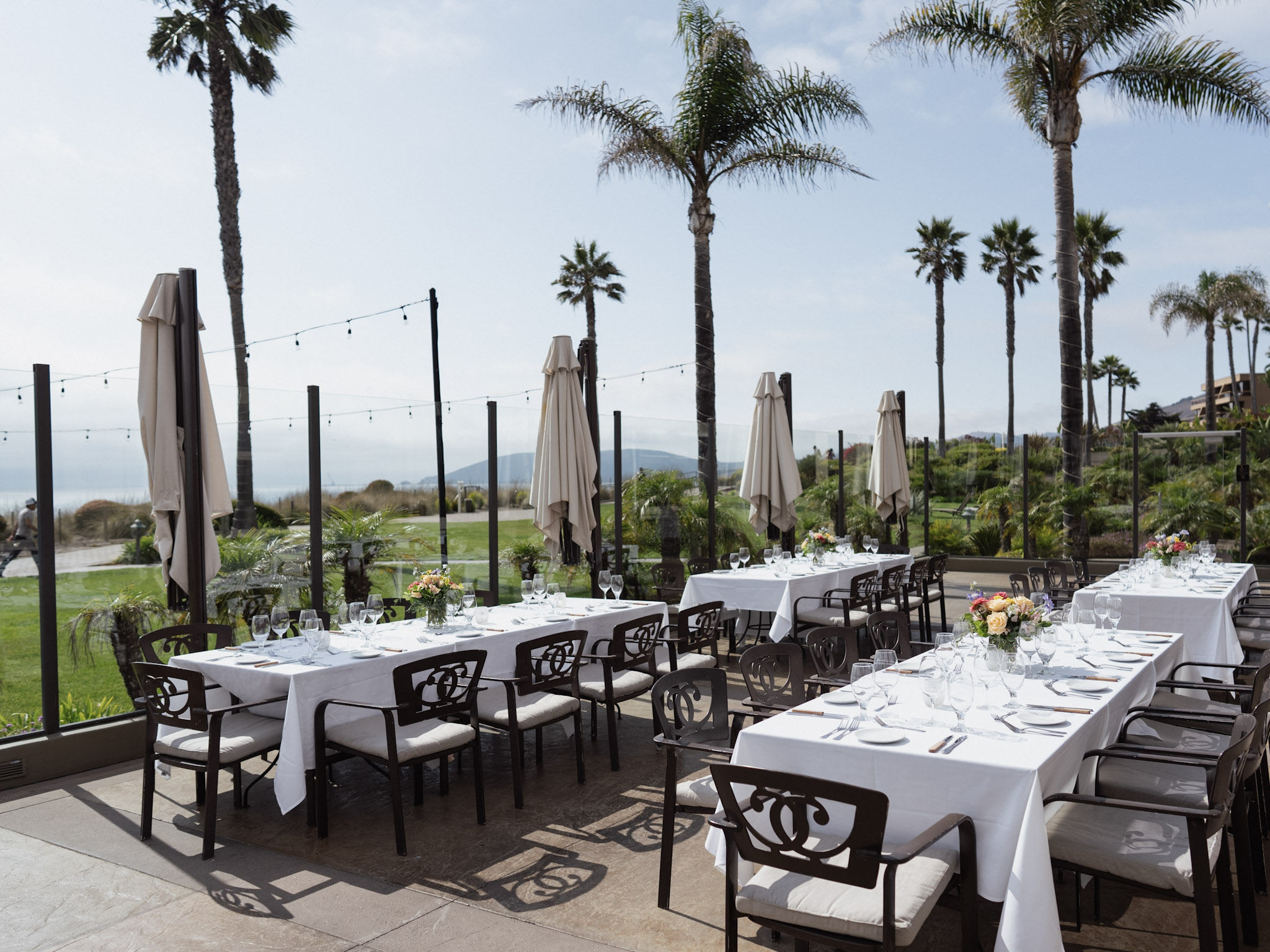 Peaceful oceanfront Celebration of Life setup at Dolphin Bay Resort & Spa in Pismo Beach featuring white linen tables, floral arrangements, palm trees, and serene coastal views.