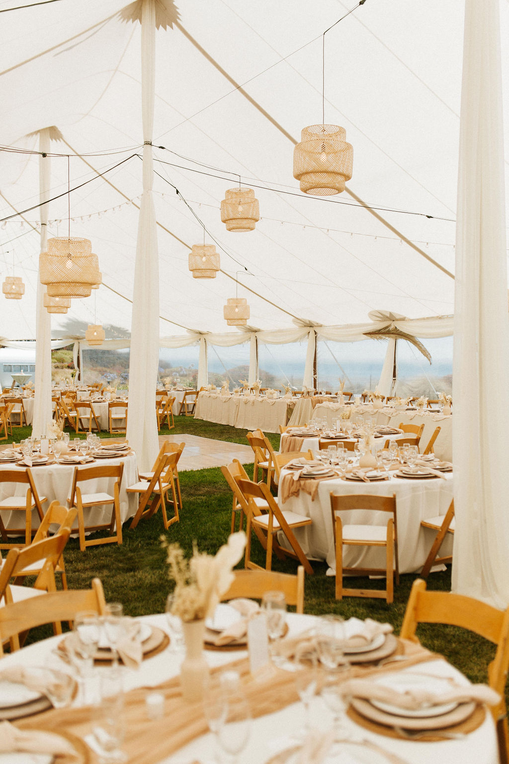 Bohemian-inspired wedding reception inside the Pacific View Tent at Dolphin Bay Resort & Spa in Pismo Beach — featuring wicker lighting, natural tones, and a single long dining table with ocean views.