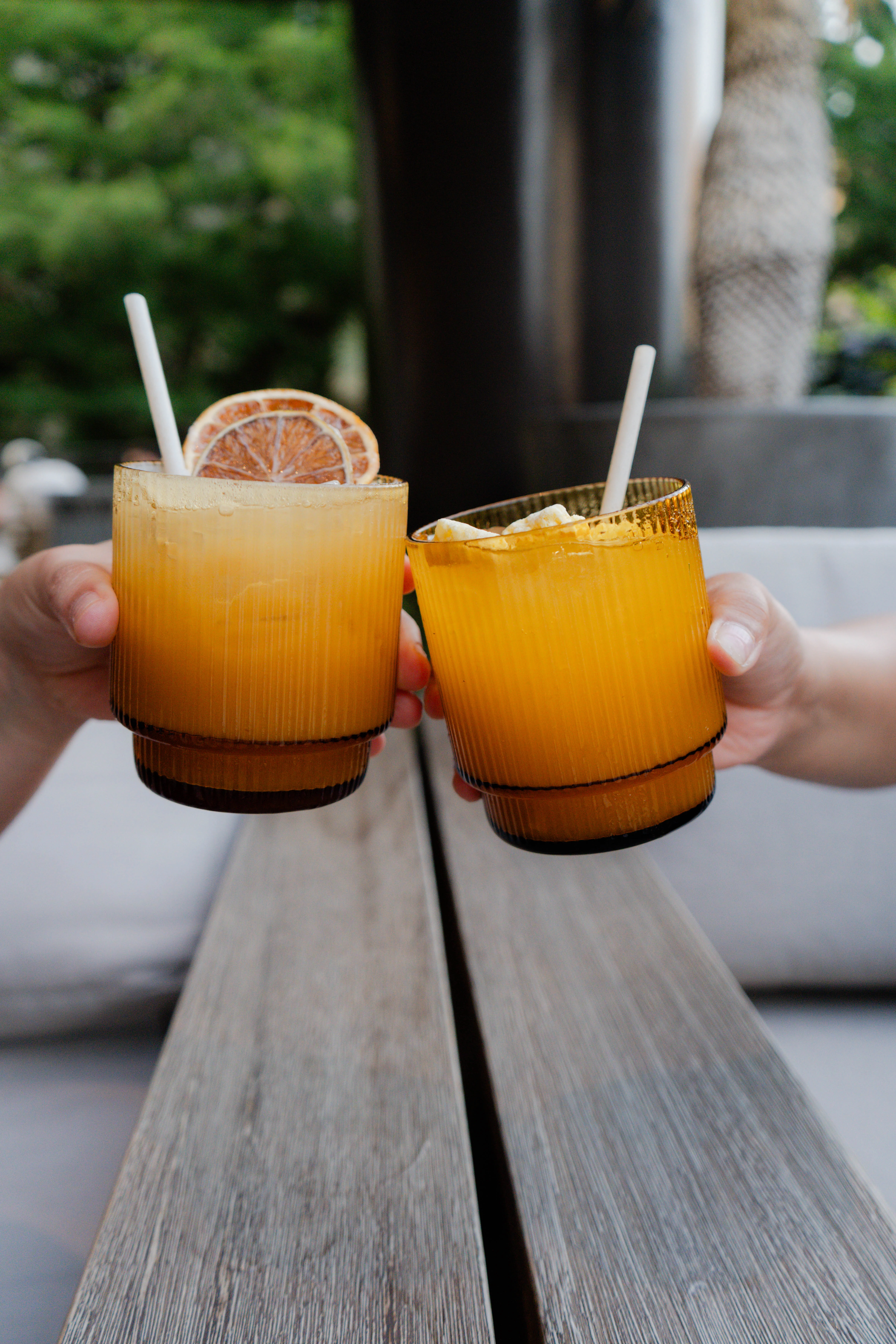 two hands holding glasses with orange liquid and a slice of orange