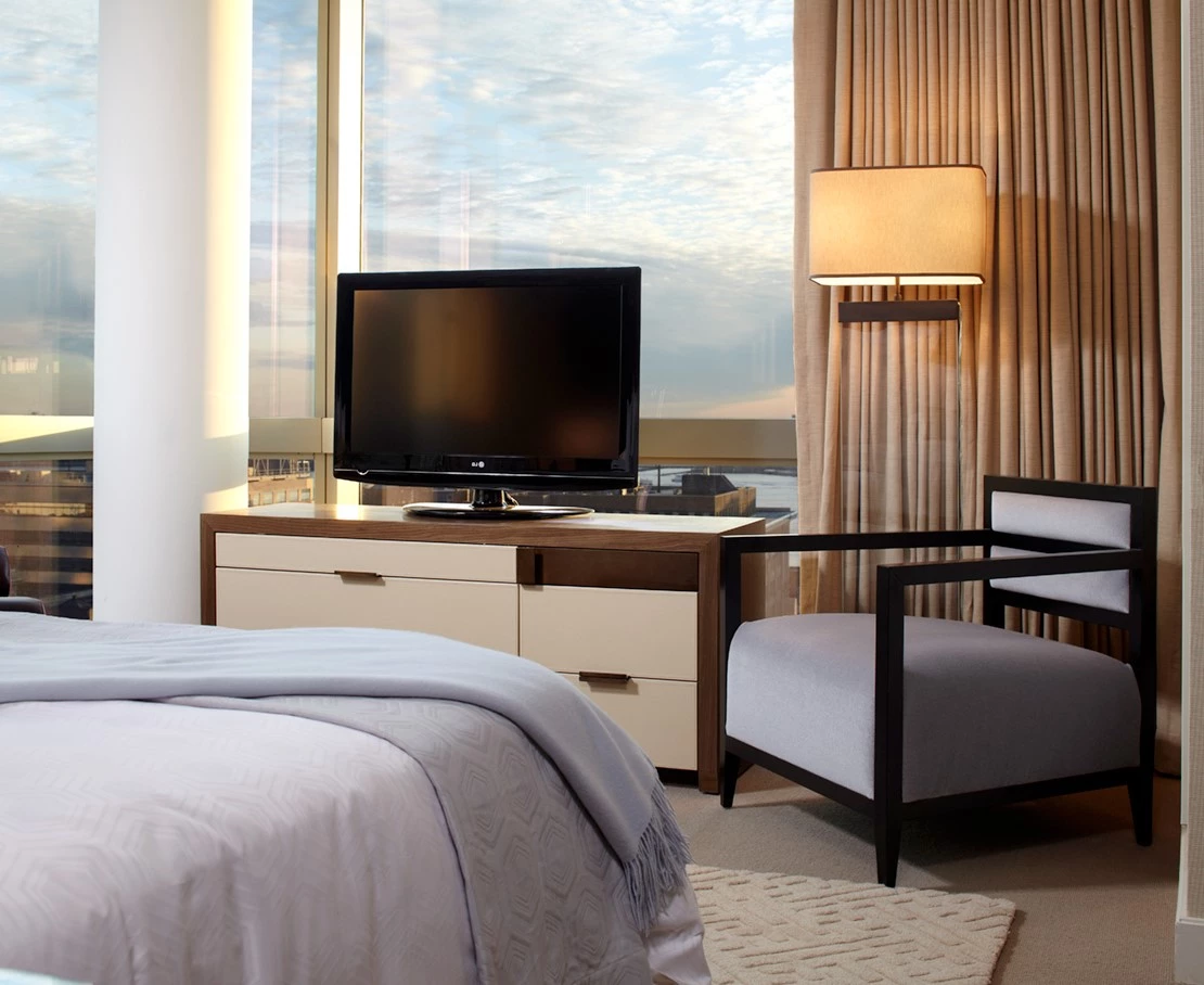 A hotel room with a bed, TV, chair, and lamp in front of a large window showing a city view.