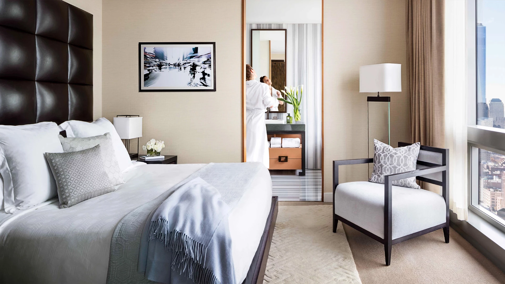 A hotel room with a large bed, chair, and window showing a city view